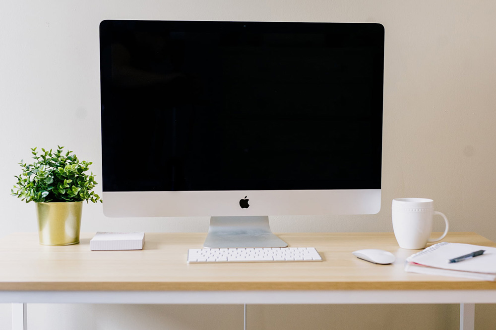 Home office desk setup, showing a Mac, keyboard and mouse, along with a mug and notepad