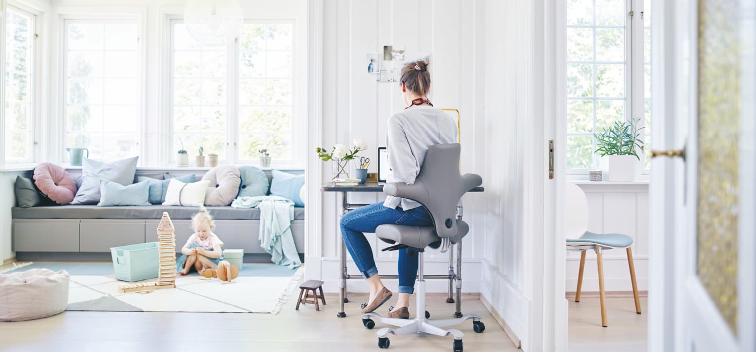 Woman working at her living room office desk while a child plays in the background