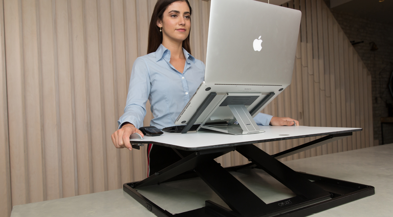 Woman raising her Oploft platform higher, gripping the side handles whilst laptop on platform