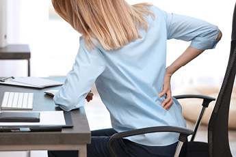 Image of a woman holding her lower back, in obvious discomfort, while sitting on an office chair
