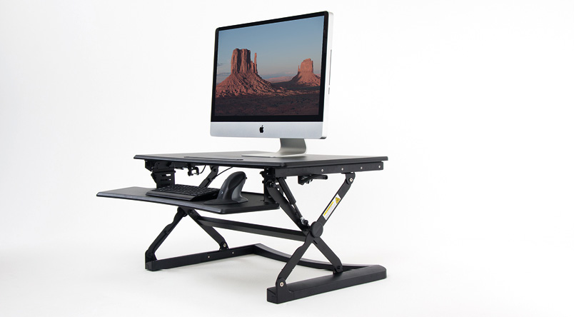 Front angle view of the black DeskRite 100 Sit-Stand Platform, showing it raised in the standing position, with a separate keyboard and mouse