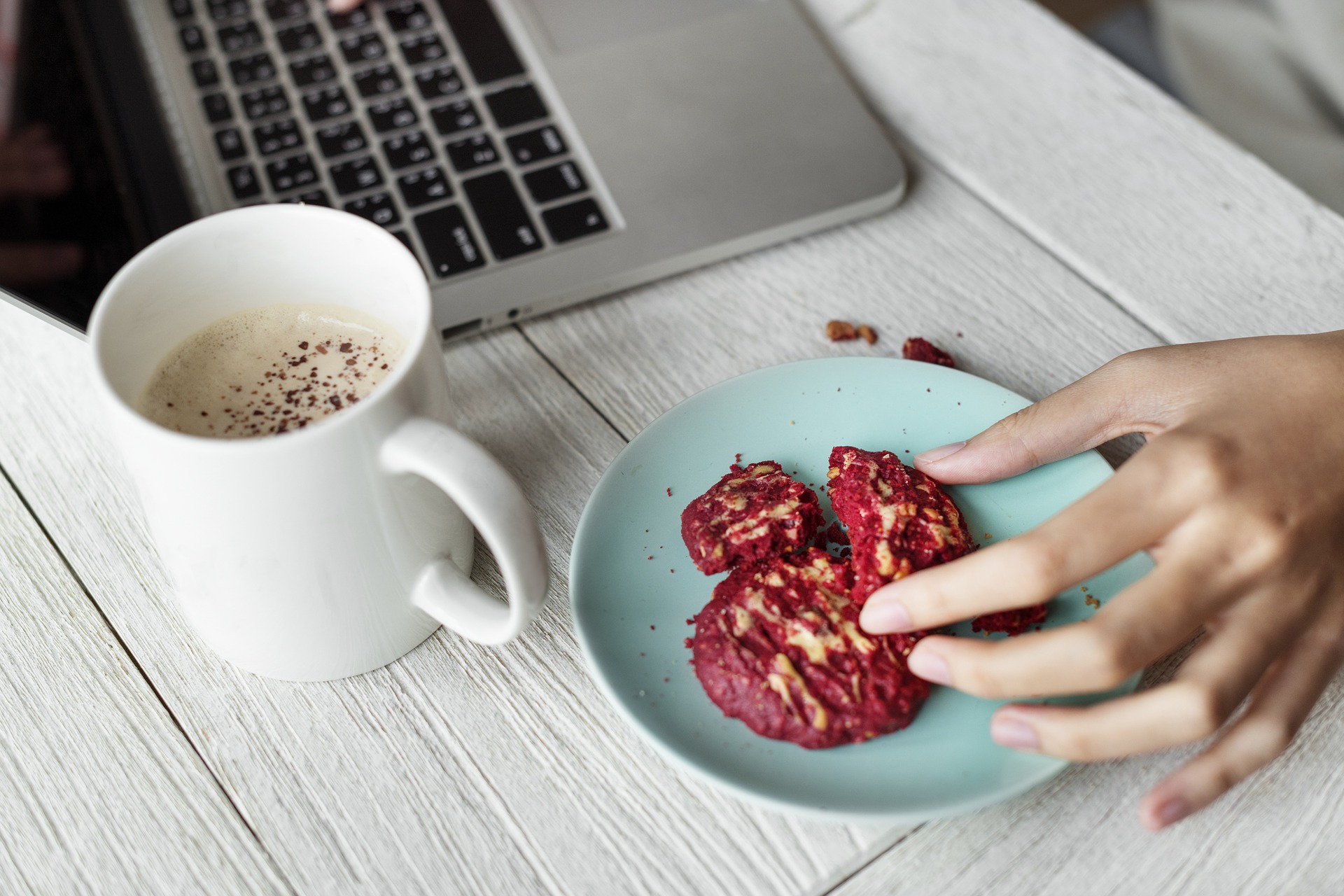 A hand reaching for a cookie on a plate, next to a mug of coffee and a laptop